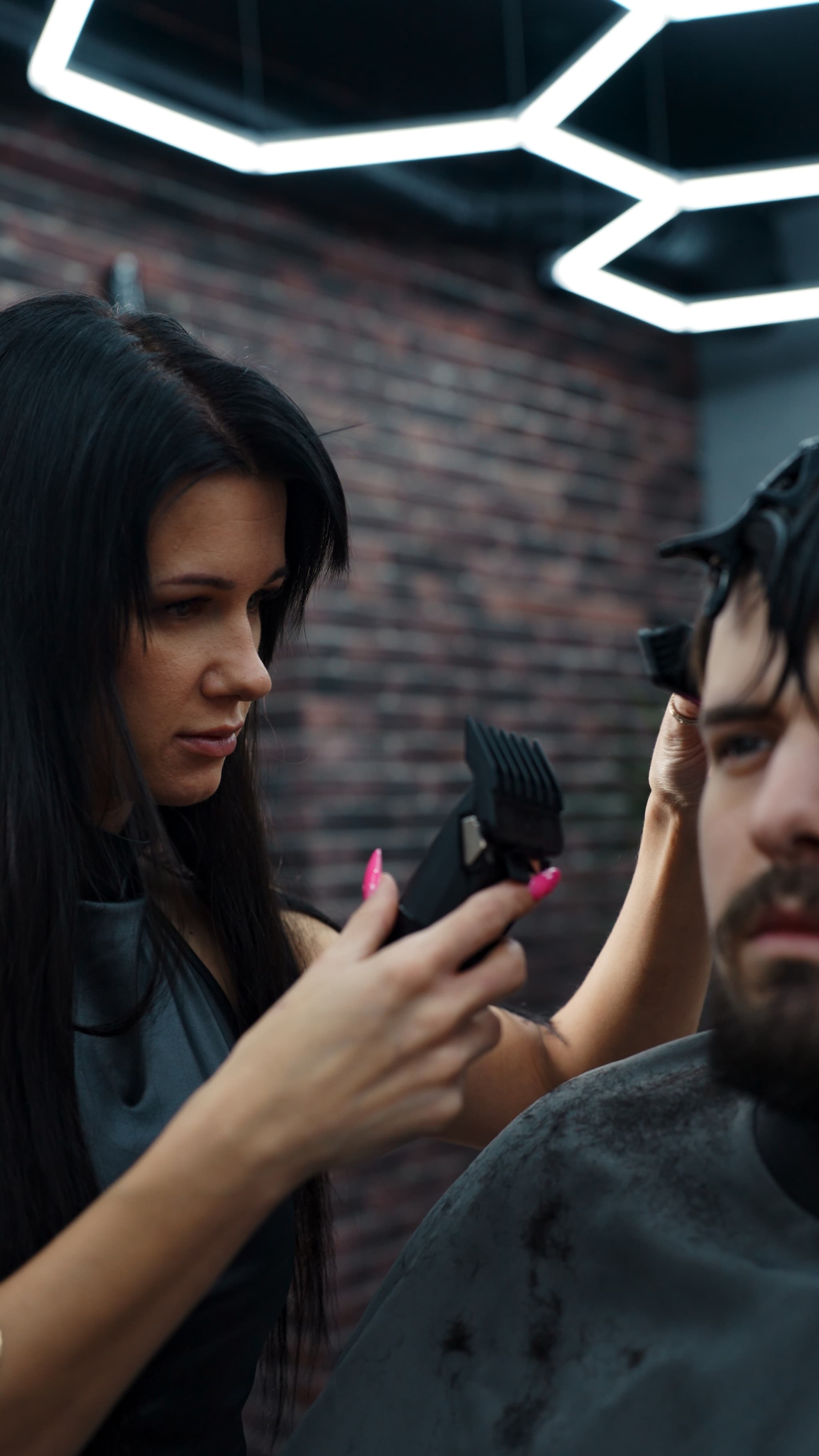 Professional female barber with long dark hair using clippers to style a male client's hair in a loft-style barbershop.